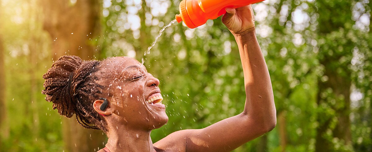 Femme se rafraîchissant en versant de l’eau sur sa tête tout en portant des écouteurs de sport Philips à l'extérieur